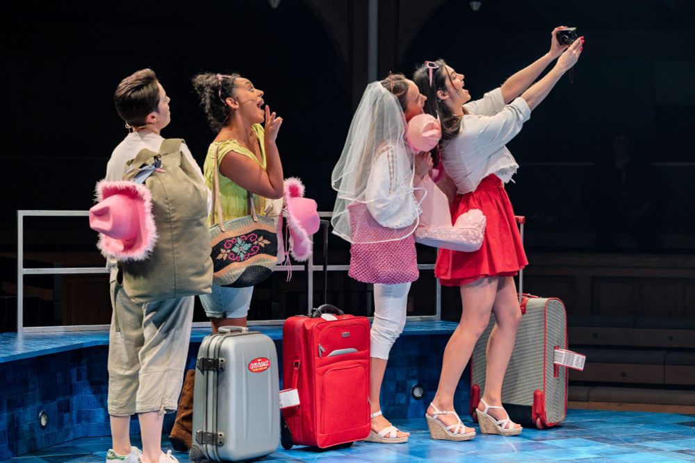 A group of women standing with suitcases, while one of them takes a selfie of the group. 