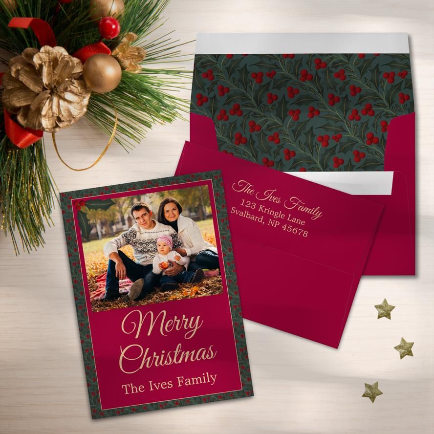 A Merry Christmas card featuring a family photo. The border is green holly leaves and berries.  Behind it are two matching red envelopes. one shows the same holly leaves and berries pattern on the inside of the envelope, the other shows a return address printed on the back in a gold color.