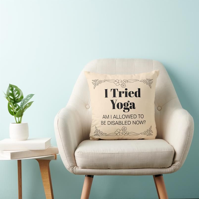 A light peach throw pillow on a white chair. On the pillow is a floral border and text which reads "I tried yoga, am I allowed to be disabled now?"