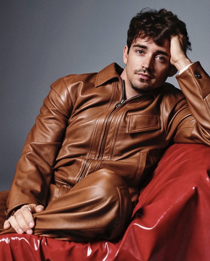 Charles Leclerc wearing a leather outfit and sitting on a red chair. He is leaning his head on his hand.