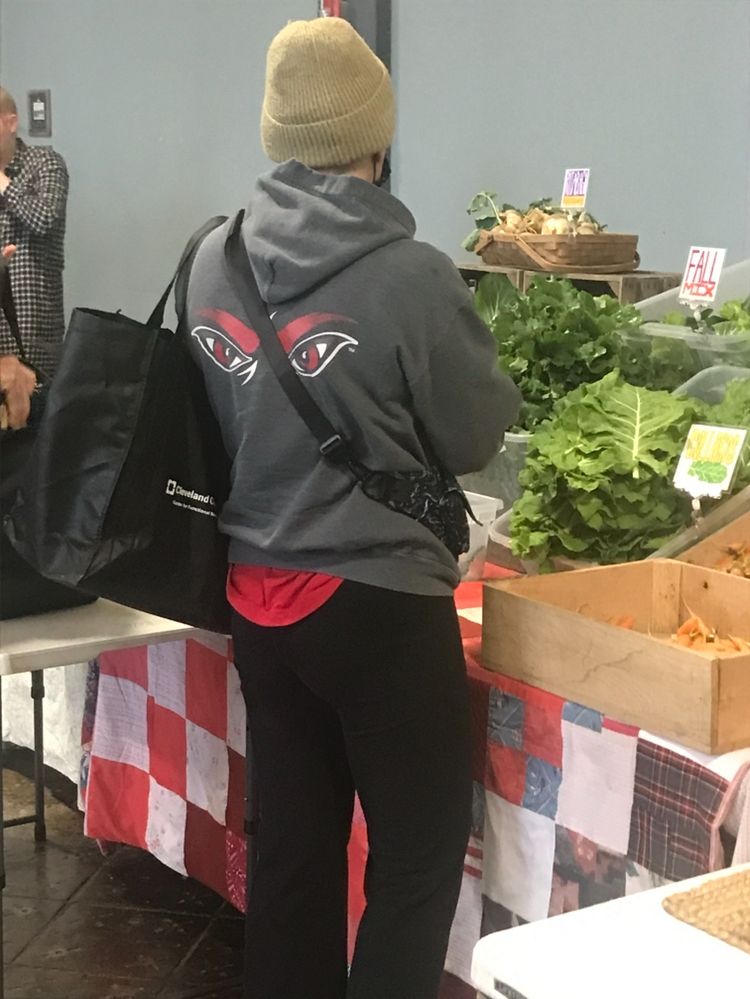 a young woman at the farmer’s market is wearing a local sports team hoodie with angry eyes silkscreened across the shoulders. her red jersey underneath is shown hanging outside the hoodie adding to the illusion of a angry monster sticking his tongue out at you.