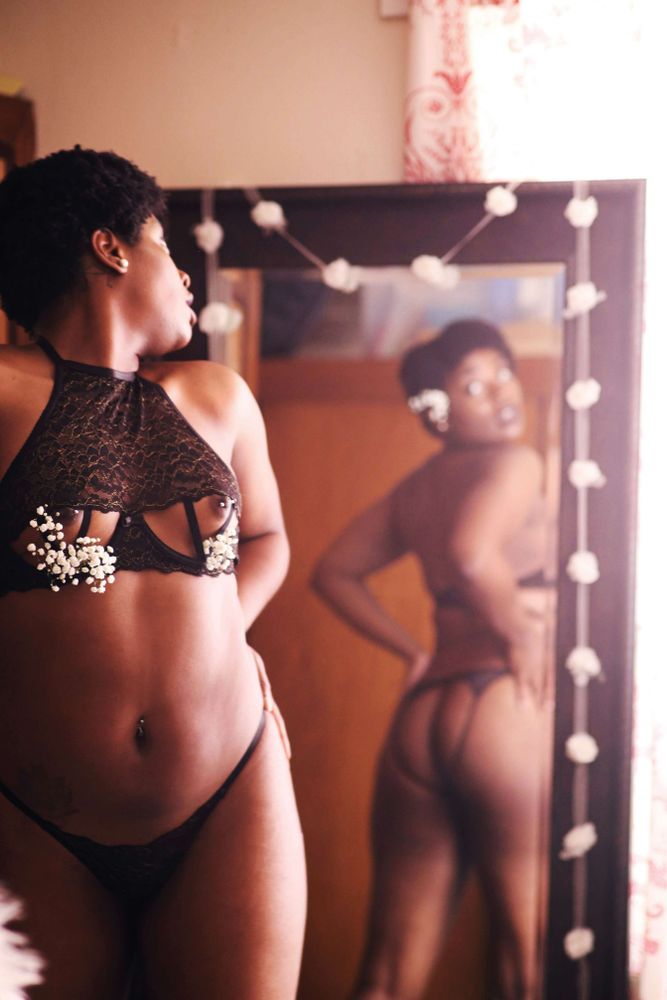 Kelly K, a Black woman with natural hair, wears a black lingerie set looking back over her shoulder in a mirror with white flowers in her hair and under the cups of her bra