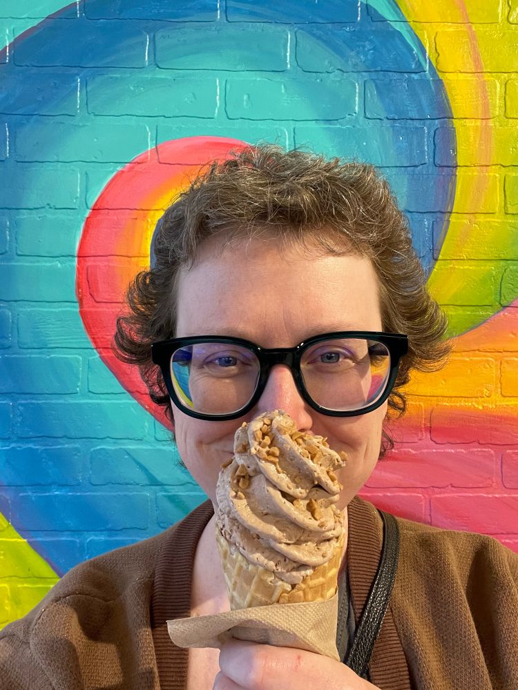 Andrea poses in front of a rainbow wall holding a chocolate ice cream cone. 