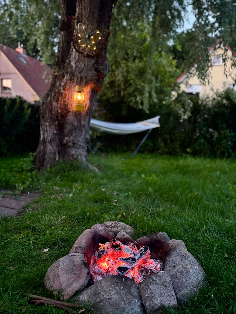Eine gemütliche Hinterhofszene mit einer Steinfeuerstelle mit leuchtenden Glut. Ein Baum mit einer Laterne und einem dekorativen Schmetterling hängt in der Nähe. Im Hintergrund ist eine Hängematte aufgestellt, und ein Haus ist im Grünen sichtbar.