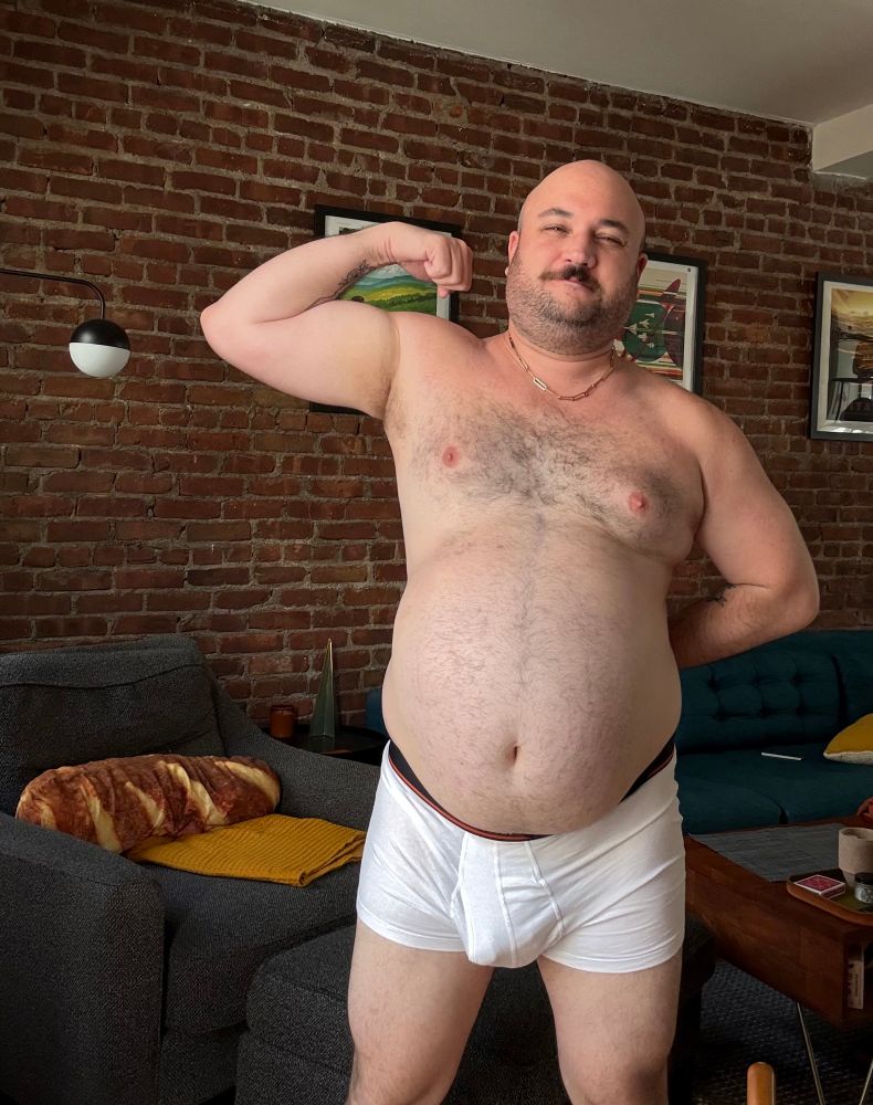flexing my bicep in front of a brick wall with a smirk on my face. My other arm is behind my back. I’m wearing nothing but a chain and white trunks through which you can see my semi erect dick