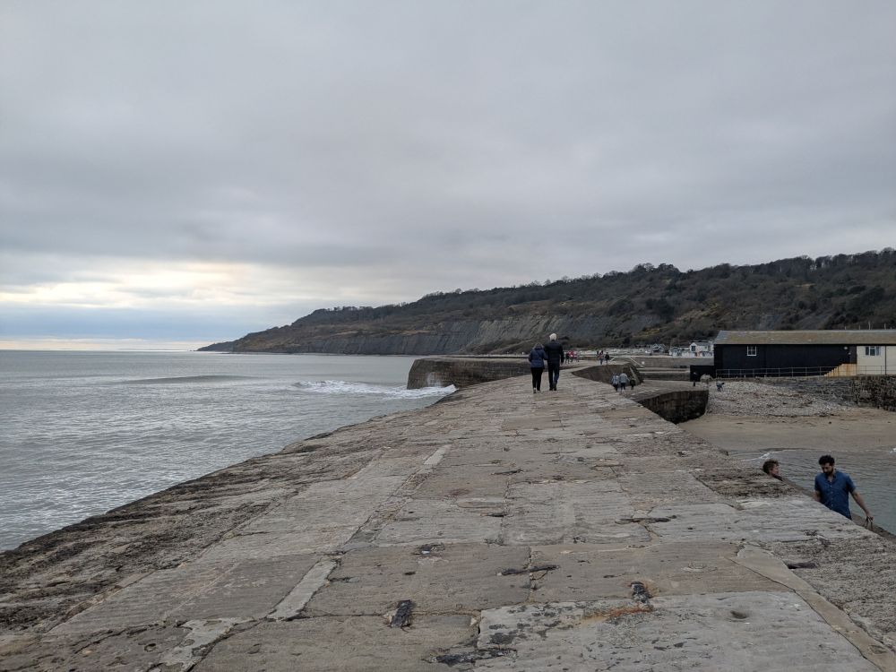 a narrow harbour wall, made of stone, with what look like large metal staples in some places.  The picture was taken facing the coast from the walkway, there is a low cliff face going down towards the water in the distance, and waves formed by rocks under the water hitting the harbour wall.