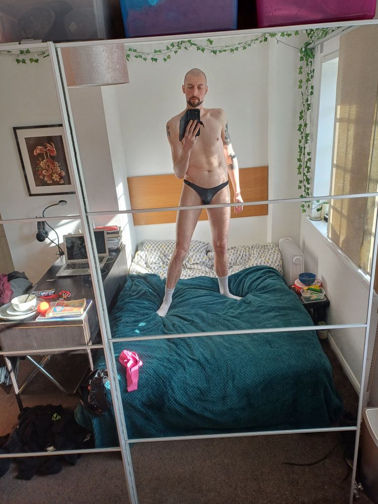 Tattooed slim guy with shaved head standing on the bed wearing a thong and white socks in the sun.