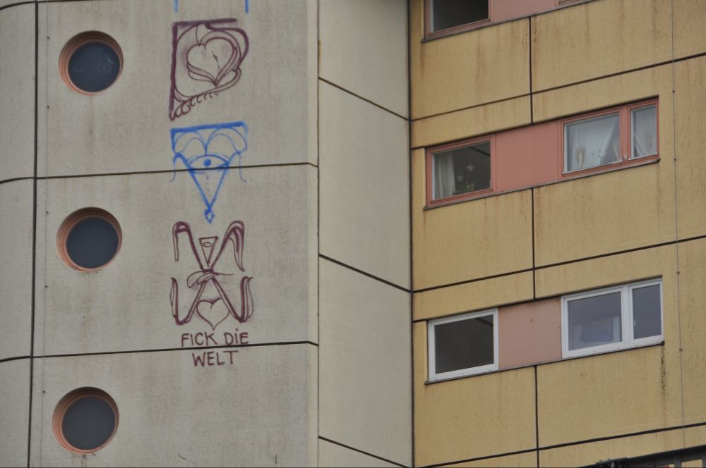 nahaufnahme der grafitti-zeichen aufm wohnblock aus den vorherigen bildern. rechts fenster, links drei runde fenster. unter den grafitti-zeichen steht klein "fick die welt"