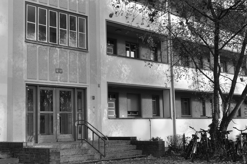 s/w. Ein mehrstöckiges Wohnhaus. Man sieht zwei Stockwerke, links der Haupteingang und rechts, die balkons und überdachten offenen gänge vor den Wohnungen.