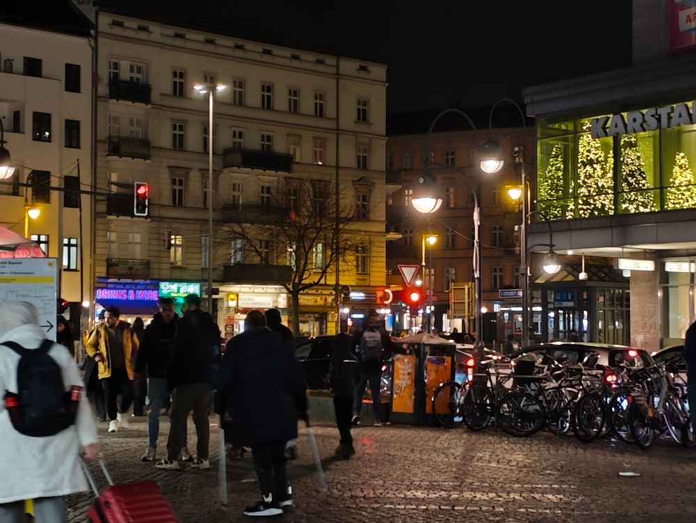 nacht. HerrmanPlatz. man sieht Passant*innen und rechts der Karstadt mit Weihnachtsbäumen beleuchtet im ersten Stock und links einen Altbauwohnhaus