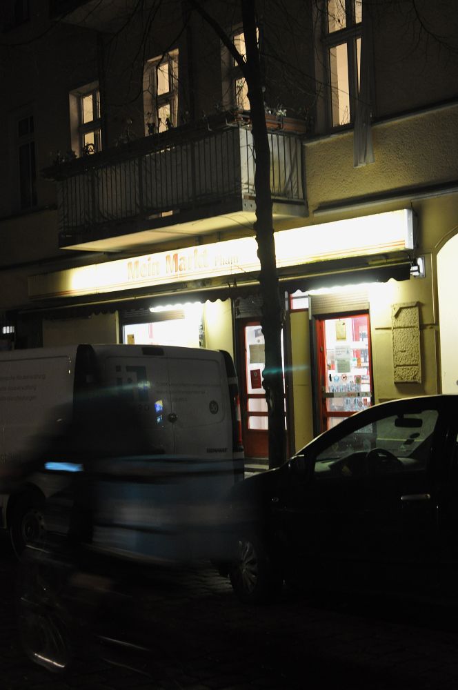 wohnhaus mit erleuchteten fenstern, unten ein laden drin "meiun markt", davor parkende autos und verwischt mit bewegungsunschärfe ein voilt lieferfahrer*in