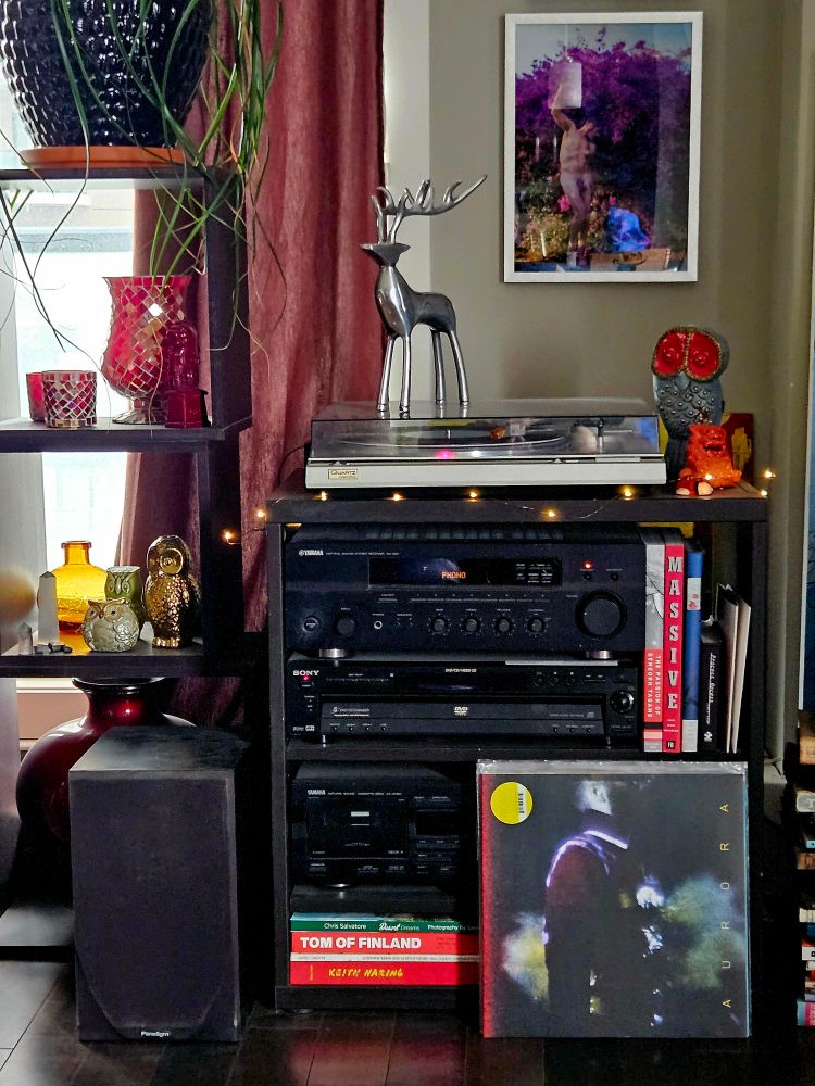 Ben Frost album Aurora propped up against my stereo shelf, which is surrounded by books and bric-a-brac. The album cover is an obscure image of a shadowed figure in right profile standing over an illuminated desk or keyboard. There is a haze of smoke. The album title runs up the right hand side of the cover. A small yellow circle with the artist's name, album title and barcode is in the upper left.