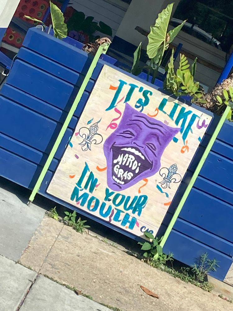 Purple Mardi Gras mask on a sign that says It’s like Mardi Gras in Your Mouth 