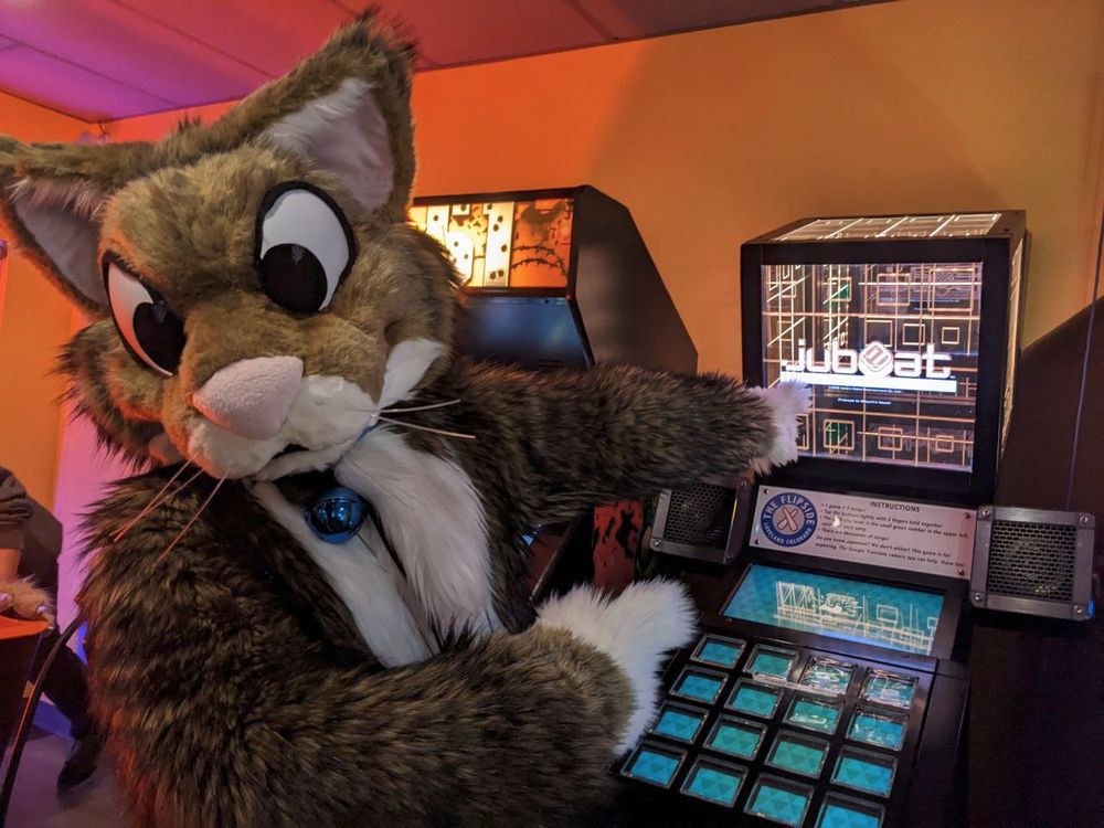 A brown and white cartoony cat with a blue bell on his collar is in an arcade. He's excitedly pointing at a Jubeat Festo machine.