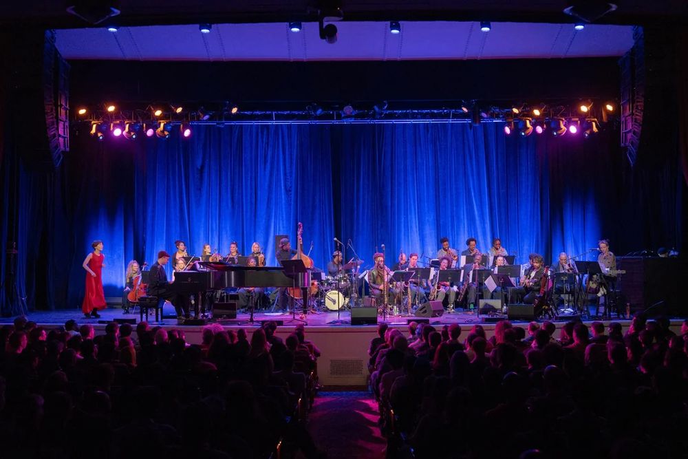 A jazz band and an all-women string ensemble is assembled on stage with composer Yoko Kanno. The curtain behind the stage is lit with blue stage lighting, and the audience in the foreground is silhouetted waiting for the show.