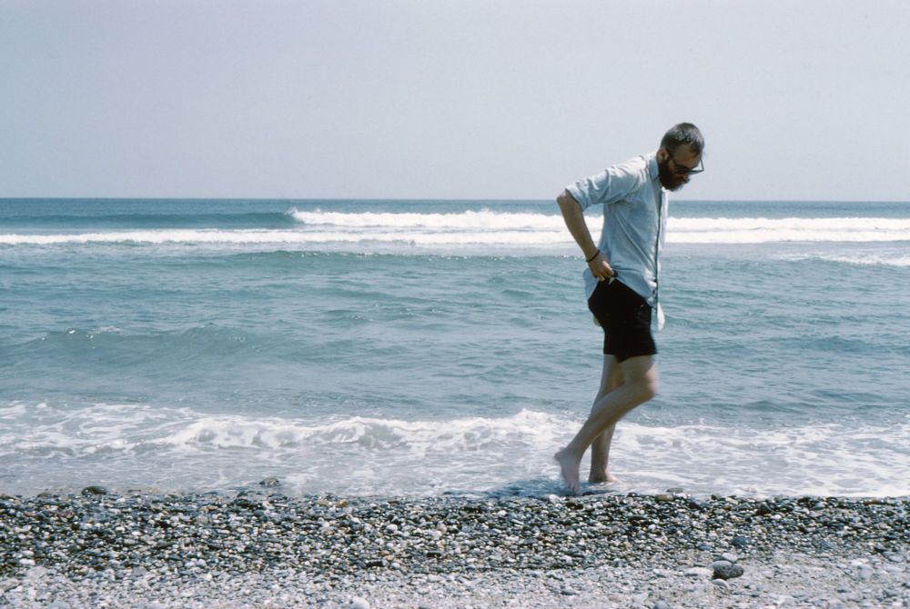 A photograph of a 36-year-old Edward walking in the water at Sandy Neck. He wears black shorts, sunglasses, and a white button-down. He is barefoot. Photograph by Eleanor Garvey. From the Collection of Ken Morton.