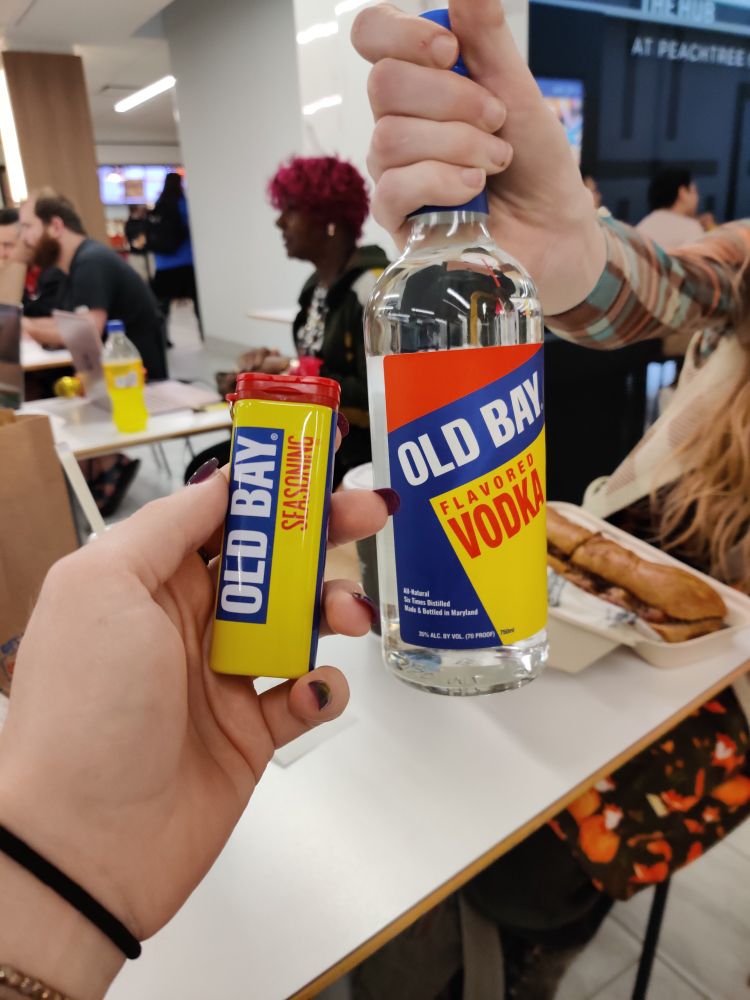 A picture of two hands holding up an old bay keychain shaker and a container of old bay flavored vodka.