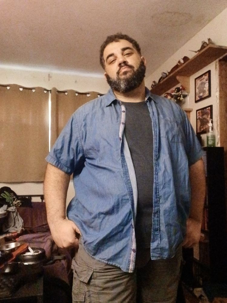 Scott is standing in his bedroom wearing a pair of slate gray cargo shorts, a dark gray t-shirt, and a denim blue open button shirt. 