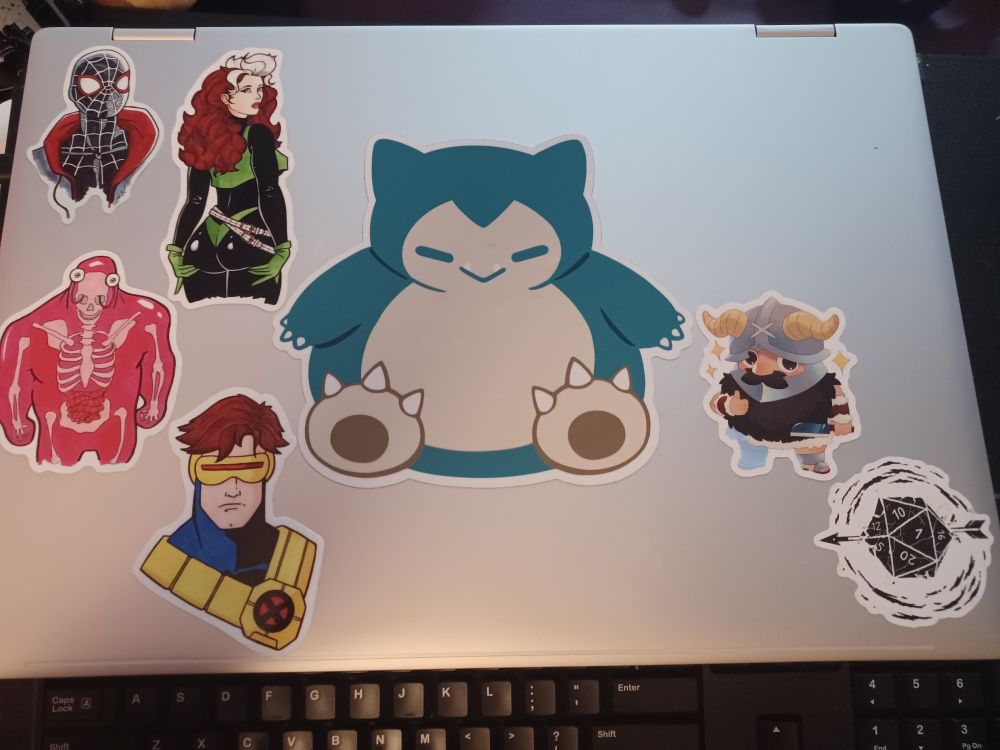 A shot of a silver HP Envy laptop cover. The HP logo is covered by a medum sized big adorable Snorlax sticker. On the left side there is a Miles Morales headshot sticker, a old school X-Men Rogue costume head to waist sticker (where she is facing away and cupping her butt cheeks while looking back at us), a head to waist X-Men Glob Herman sticker (We stan Glob!), and a headshot 90s era X-Men Cyclops sticker. On the right currently is a D20 rpg die with an arrow through it and some animated black lines encircling it as well as a Senshi from Delicious In Dungeon adorable sticker with some sparkling stars around him. 