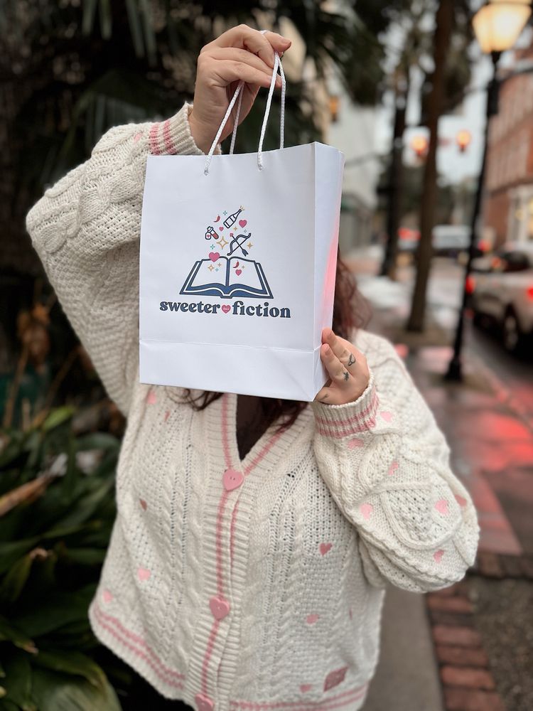 A girl standing in a white Taylor Swift Lover cardigan with pink details holding a shopping bag in front of her face. The shopping bag reads “sweeter than fiction” and has a book shaped logo 