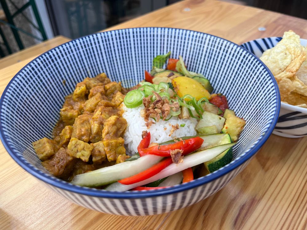 A ceramic striped blue and while bowl with tempeh rendang, rice and vegetables.
