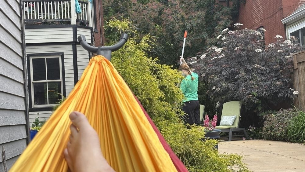 My toes are visible in the corner of a yellow & maroon hammock. In the background, partially obscured by a green bush, my teenage daughter can be seen vacuuming our backyard pool. 