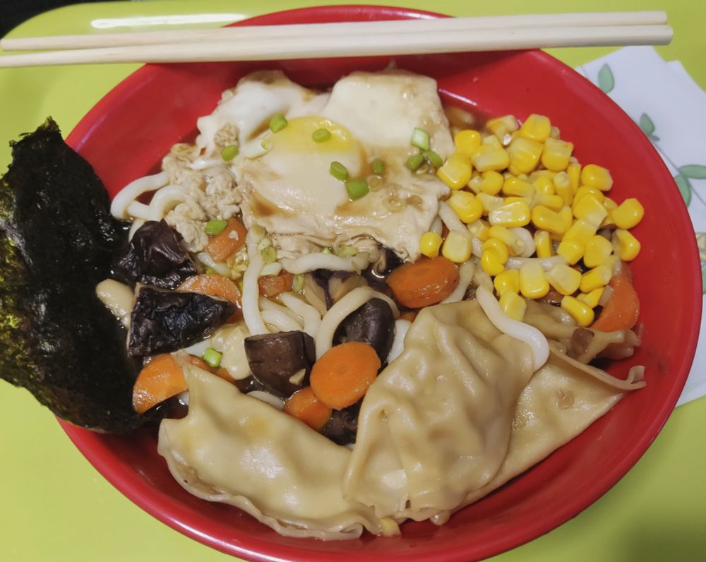 Cuenco rojo de ¿ramen? casero sobre una bandeja verde y con unos palillos reposando sobre una esquina de este.
El cuenco se compone de caldo, fideos udon (gruesos), alga kombu en un lateral, setas y zanahoria cortadas, gyozas, maíz y un huevo frito (cocido sobre caldo, más bien). Se ha adornado con un poco de verde de cebolleta cortado por encima.