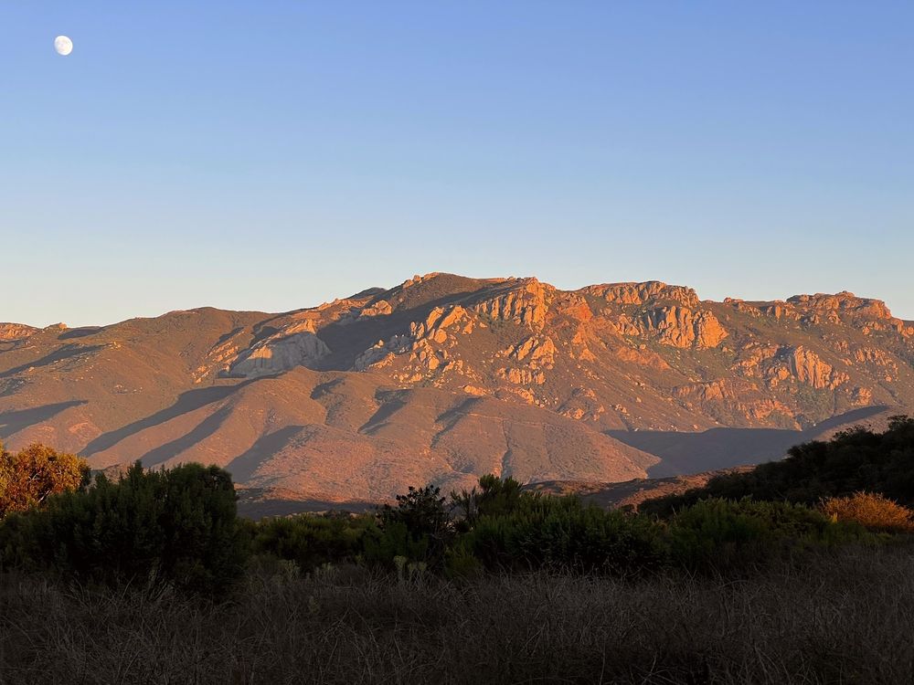 A photo of the north side of Satwiwa, a mountain in the Santa Monica mountains, with dramatic golden hour sunlight coming in from the right. There's chaparral in the foreground and a soft baby blue sky above. In the top left corner of the image, inexpertly unaligned to any thirds, you can see the moon, almost full. 