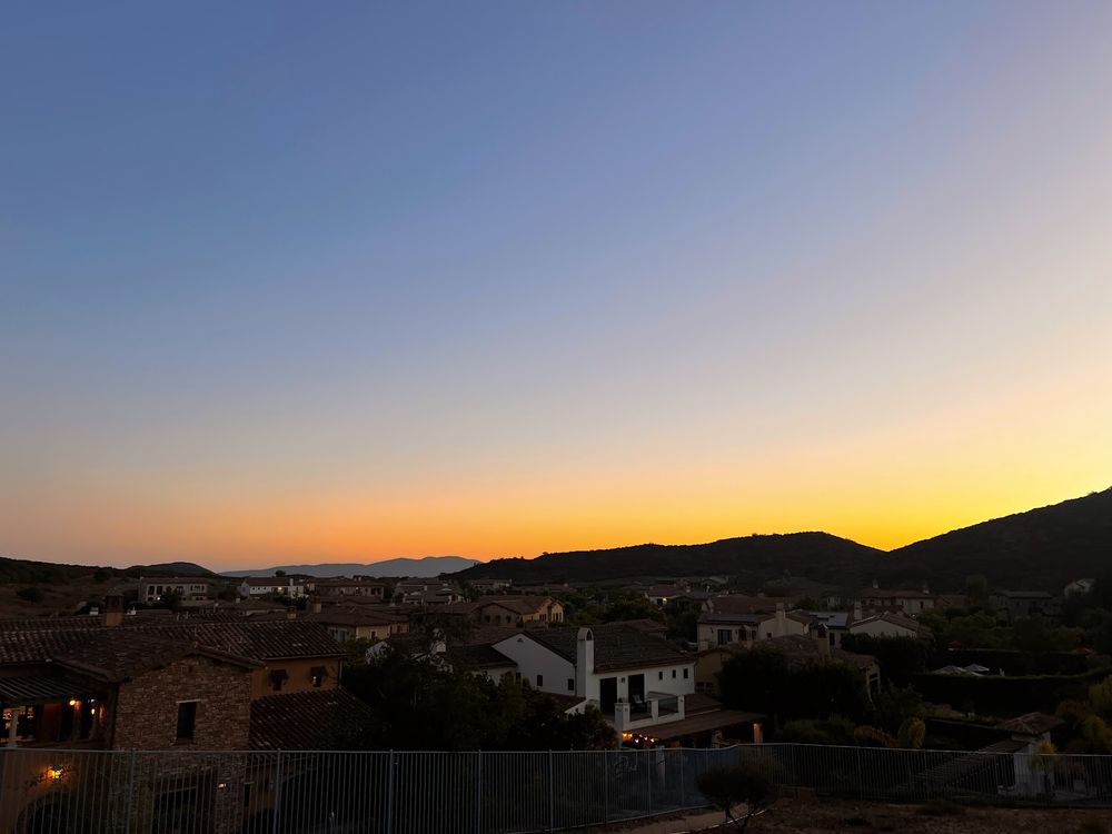 A sunset over a SoCal coastal community. The sky ranges from bright yellow on the right end of the horizon to a soft peachy color on the left end of the horizon. Above the yellows are various gradients running to a white-blue on the right and a deeper sky blue on the left. 