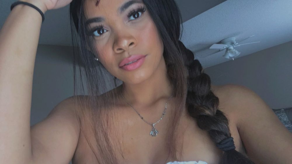A selfie of Goddess Suviina. She is facing the camera with her eyes open wide & bright. Her long eyelashes hover over her dark brown eyes. Her lips are petal pink while her cheeks are flushed with red. Her long thick hair is in a braid down the right side of her head with two loose strands in the front of her face. She is wearing a white tube top that is not visible in the frame. She has on a silver necklace with a angel heart pendant. 
