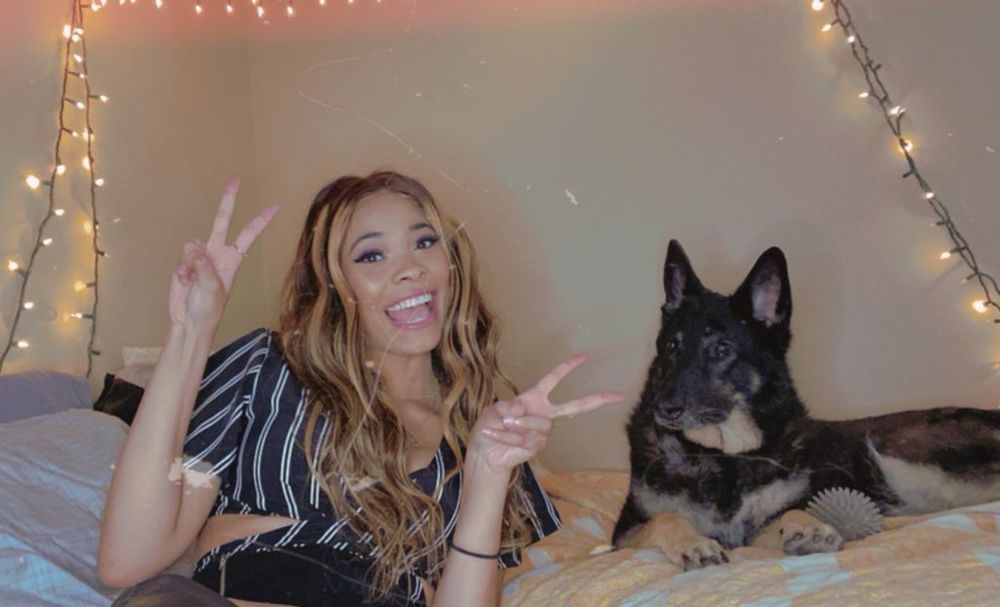 Suvii & Varsity the German Shepherd are lying on Suvii’s bed. 

Suvii is holding up peace signs. She has blonde brown hair with highlights. She is also wearing a striped black & white top. She is a Black-Biracial woman. 

Varsity is staring at Suviina while holding onto her gray ball. 
