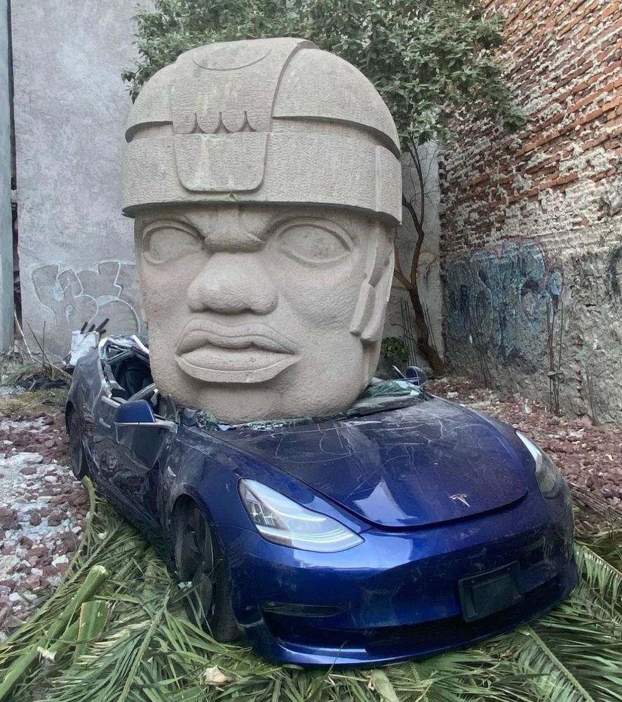 Post found on instagram from the account “chavismarmol”. We see a giant sculpted replica Olmeca head weighing approximately nine tons dropped on a midnight blue Tesla model 3, completely destroying the piece of shit car