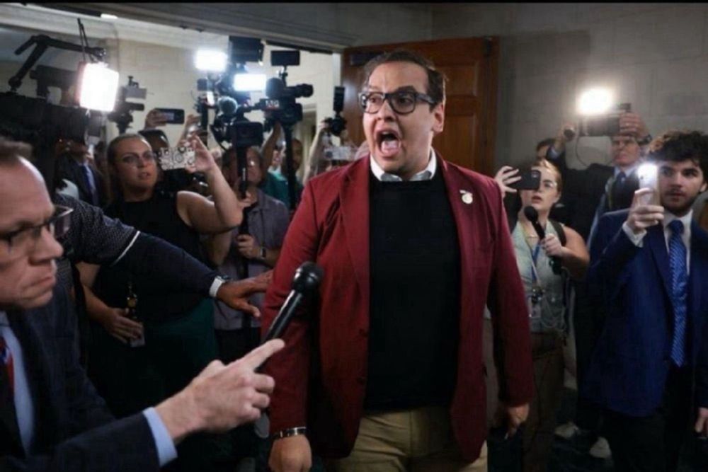 George Santos shouting into a microphone, flanked by media outlets