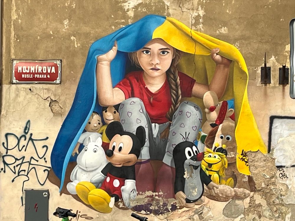 Fresco painting on a building of a young white girl with blonde braids, using the Ukrainian flag to provide shelter for herself and famous cartoon characters rendered in he size of medium sized dolls.