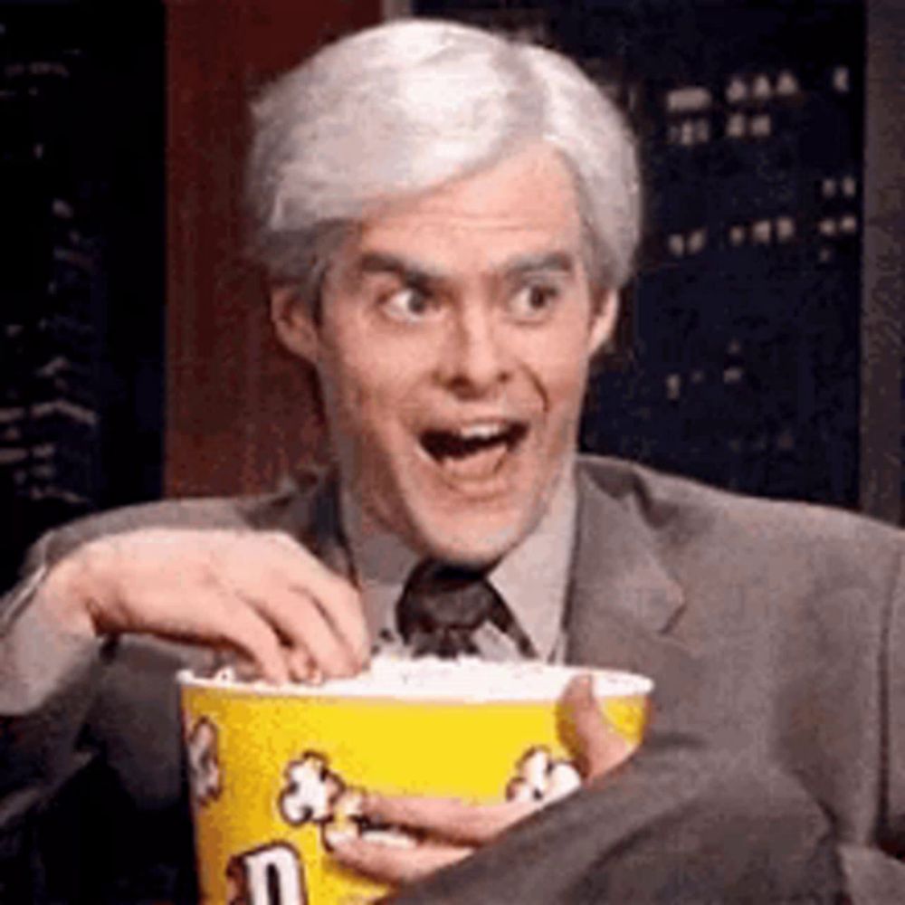 a man in a suit and tie is holding a bucket of popcorn and eating it .