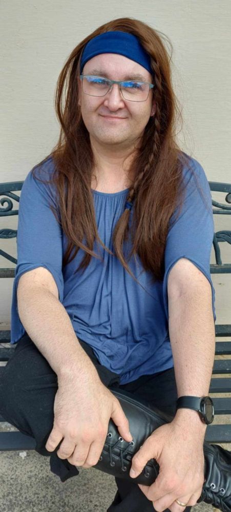 A transgender woman seated on a bench, wearing a blue blouse with a flowing design and long brown hair styled with a blue headband and braid. She is smiling warmly, wearing glasses and black boots, with hands resting on her leg.