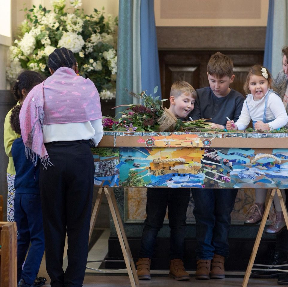 Two groups of children and an adult gather closely around a brightly decorated cardboard coffin with pens in their hands.