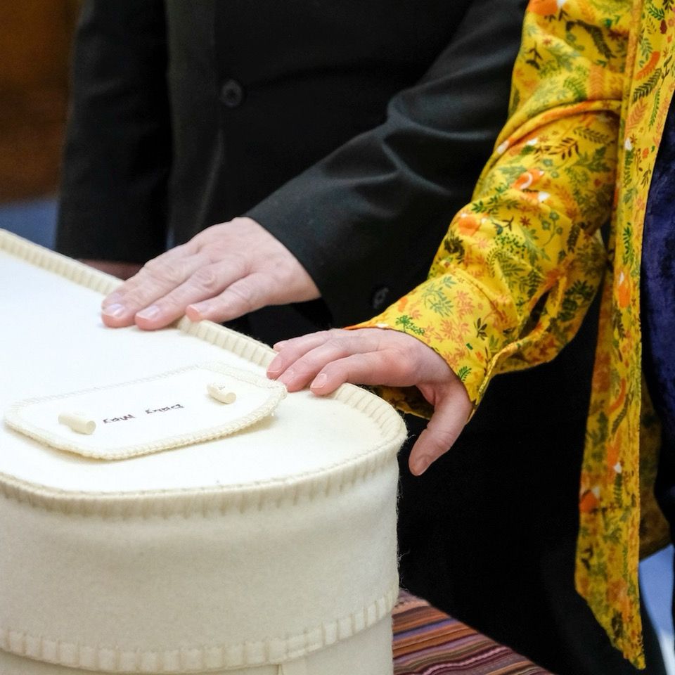 Two hands touching a small, cream coloured woollen coffin.