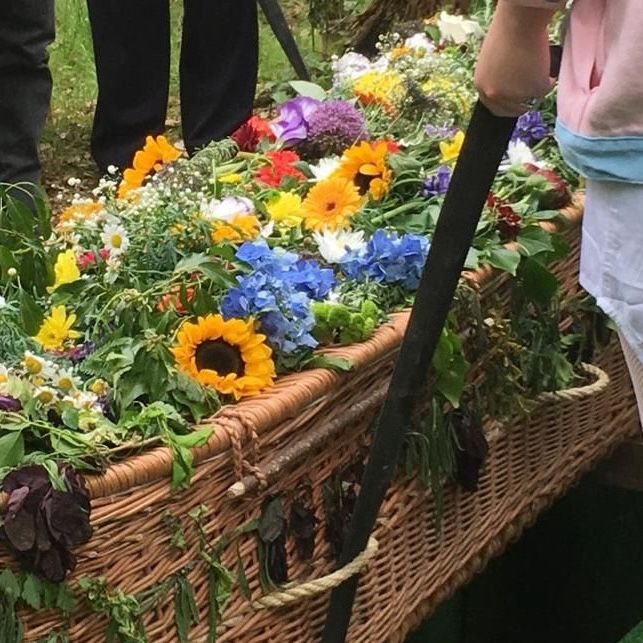 A close up shot of a young person’s hand holding a coffin strap supporting a willow coffin covered in flowers.
