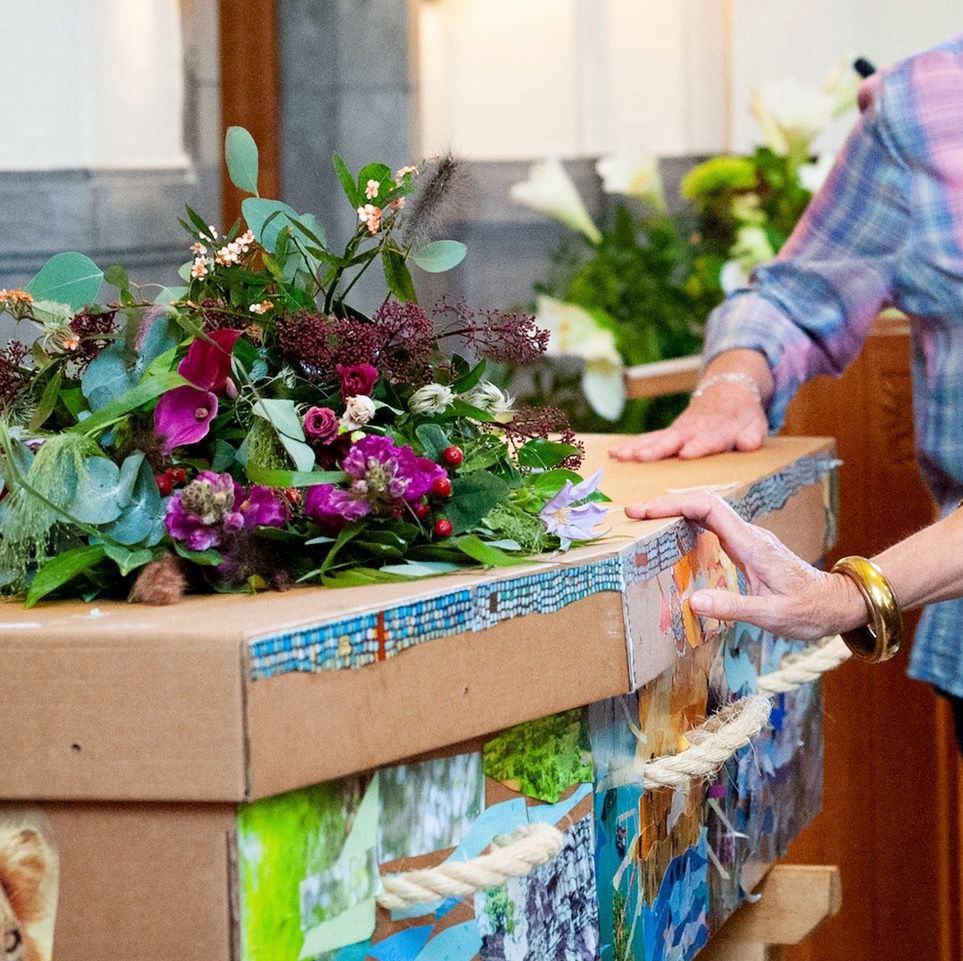Two witw women’s hands touching a cardboard coffin with rope handles, decorated with pictures, with a bouquet of wildflowers on top.