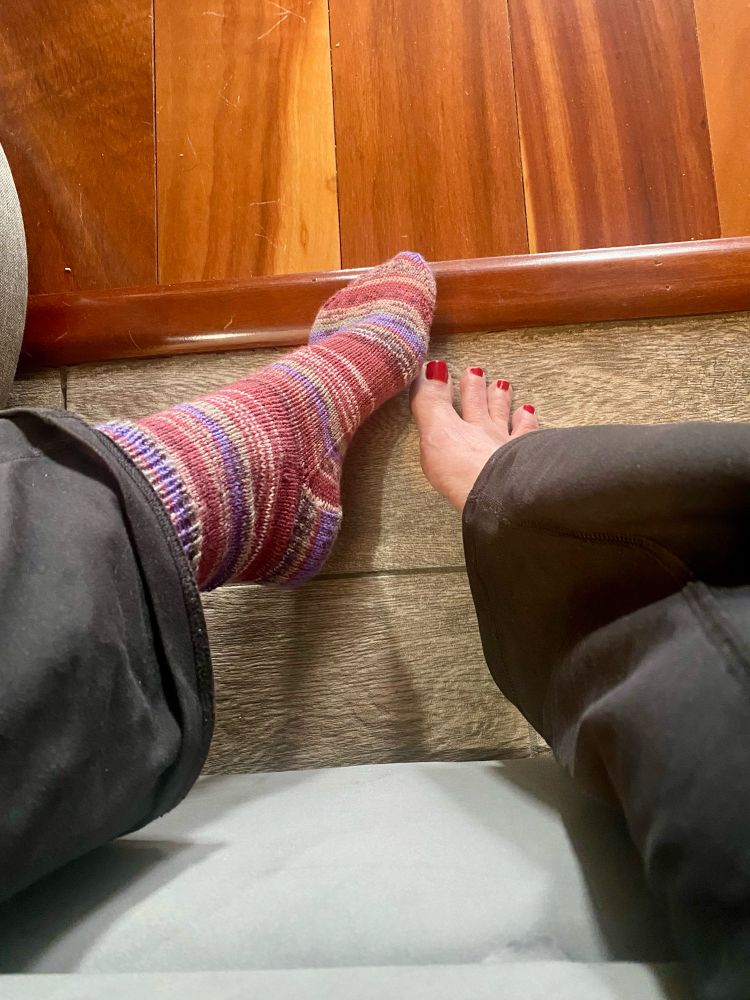 Feet on floor. Left foot in fresh off the needles knit sock, purple brown pink variegated yarn, right foot bare.