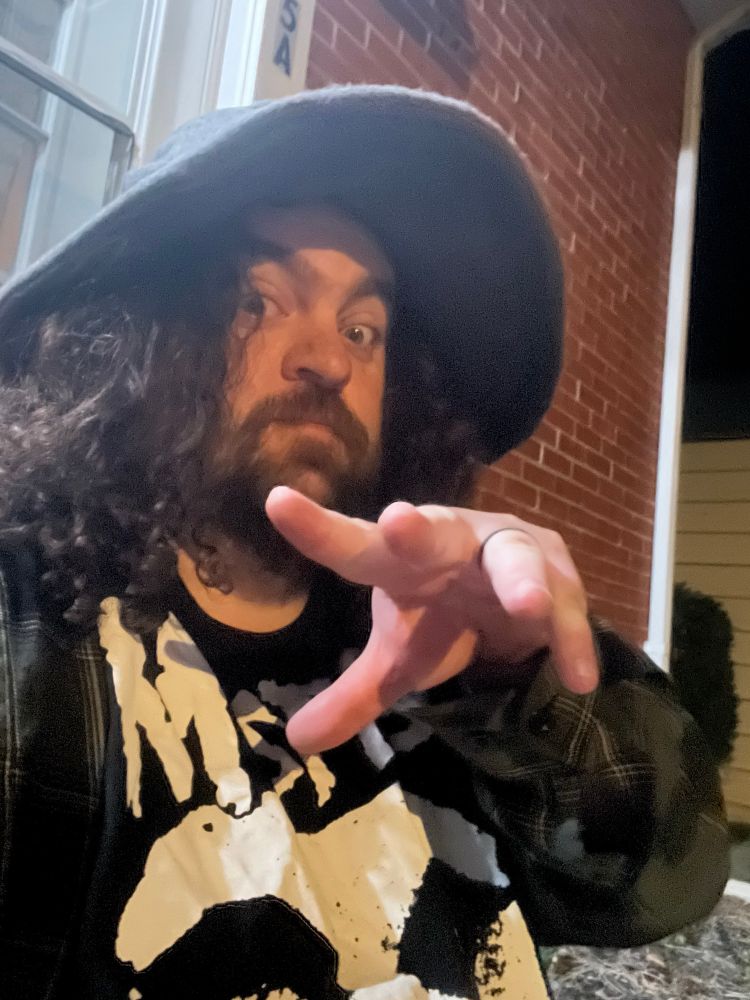 A long haired man with a beard in a wizard’s hat, a green flannel, and a Misfits t-shirt stares at the camera with a mysterious look in his eye, his left hand appearing to be slinging some mighty powerful sorcery, or giving jazz hands, only the pictured man and God himself knows
