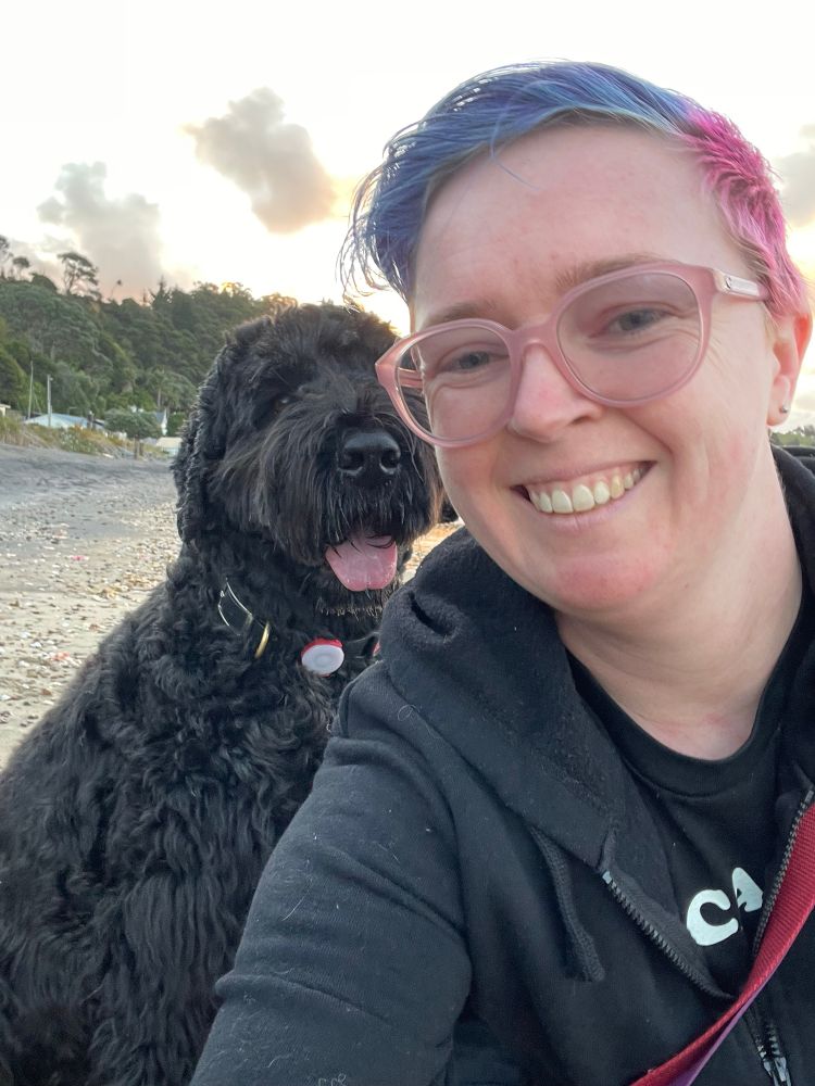 Selfie of me wearing glasses. My hair is blue on top and pink on the sides. Behind me, Gus the large shaggy black dog is sitting and looking at the camera. His large pink tongue is peaking out. 