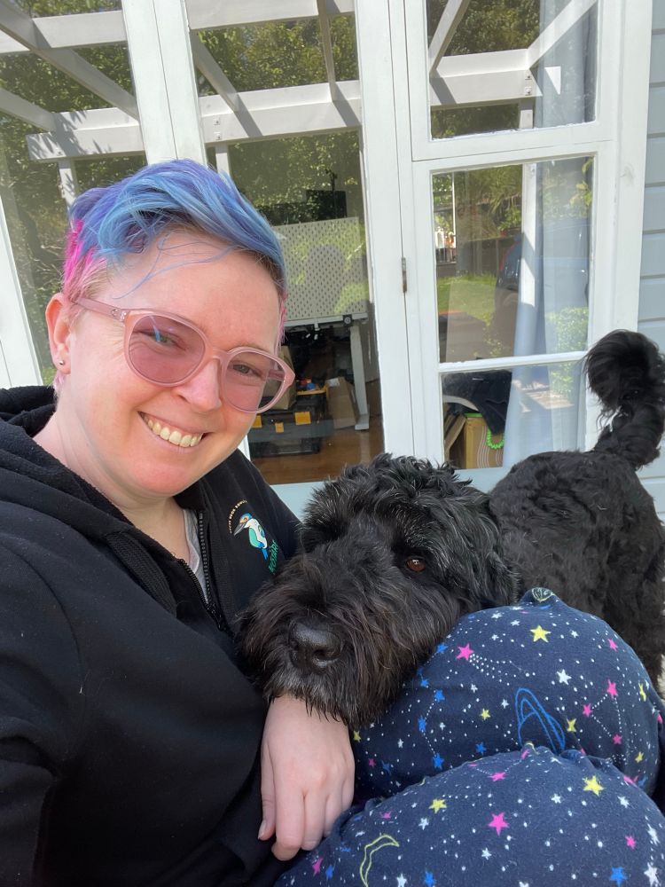 Selfie of me. Blue and pink hair, pink glasses. I’m wearing space themed navy pyjama pants and a hoodie. A large black dog is leaning his head on my thighs and gazing at the camera 