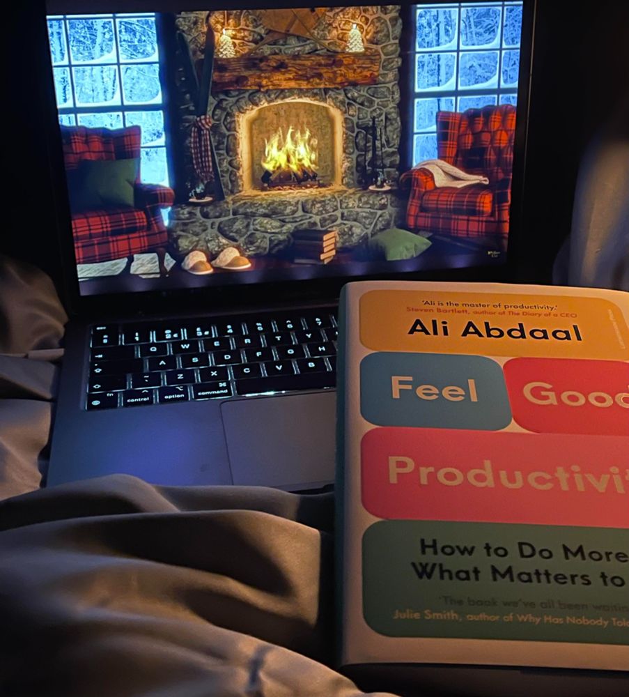 Dimly lit room with a laptop on a bed, there is a fire place on the screen and the book 'Feel Good Productivity' by Ali Abdaal on top of it.