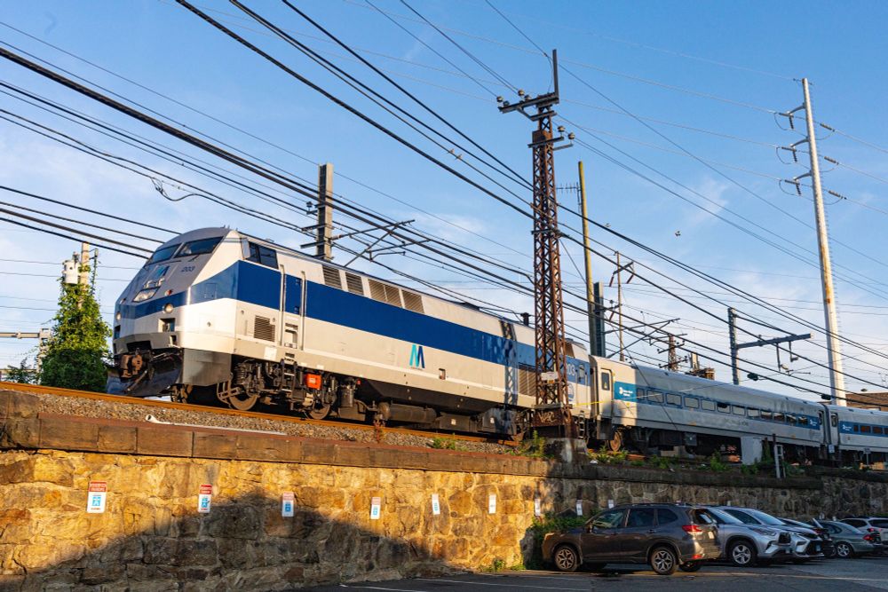 A Metro-North heritage unit locomotive running along a bridge on a sunny day, with cars parked below. 