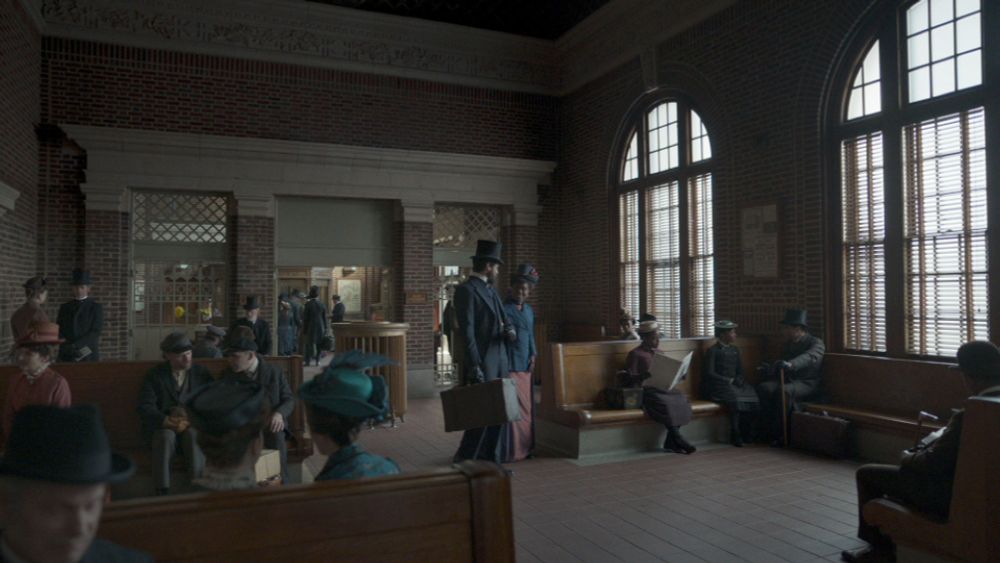 Two actors waking inside an 1880s recreation of Yonkers Station, with people seated on benches and walking around the waiting room.