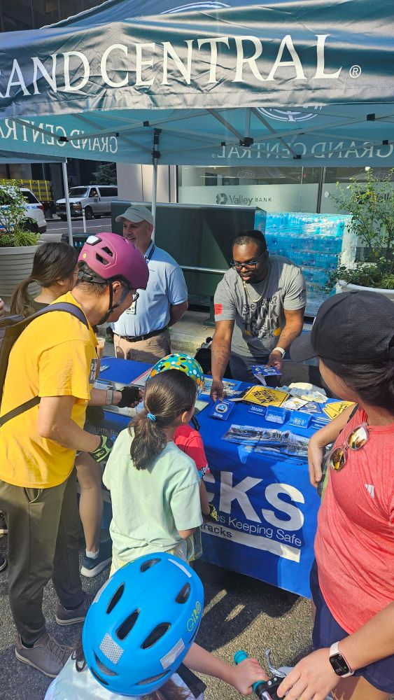 Metro-North staff interacting with people wearing helmets at the TRACKS table, containing brochure and other TRACKS safety merch, at Summer Streets.