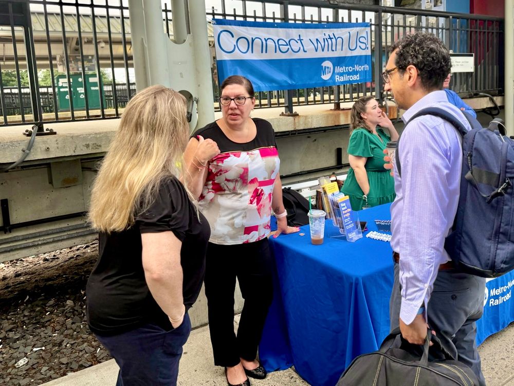 A Metro-North employee speaking with a customer at the Connect with Us table, lined with pens and brochures. 