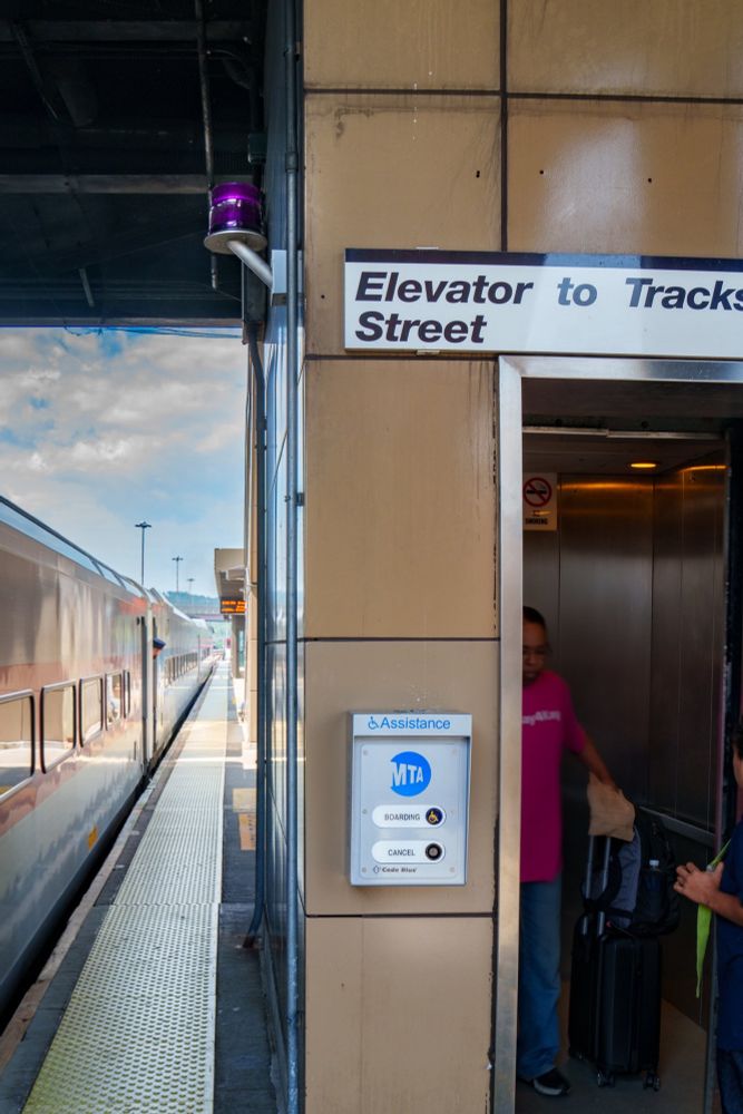 The boarding assistance button on a wall next to the elevator of Croton-Harmon station during the daytime. A train is stationed at the platform.